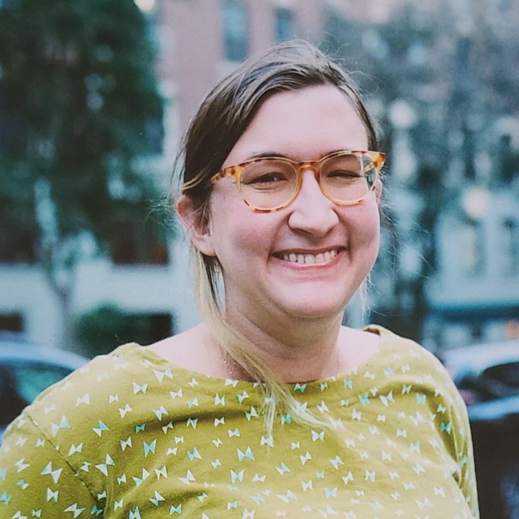 A white person with orange glasses and tied-back brown hair smiles to the camera. She wears a bright green shirt with a geometric print and stands in front of a background of blurred buildings.